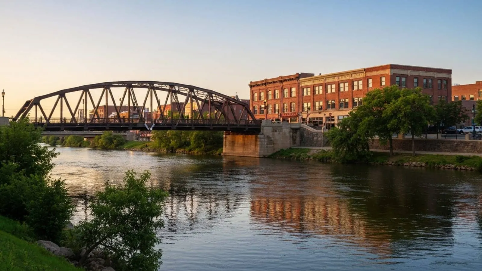 Editorial photograph of the Fox River bridges in downtown Elgin Illinois at golden hour with the historic Kimball Street truss bridge and brick downtown commercial buildings.