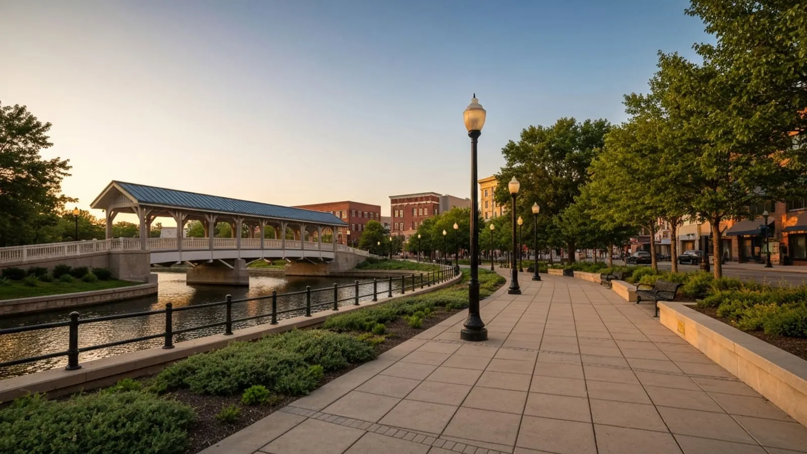 Editorial photograph of the Naperville Riverwalk along the DuPage River at golden hour with the iconic covered footbridge and the historic downtown Naperville brick streetscape visible in the distance.