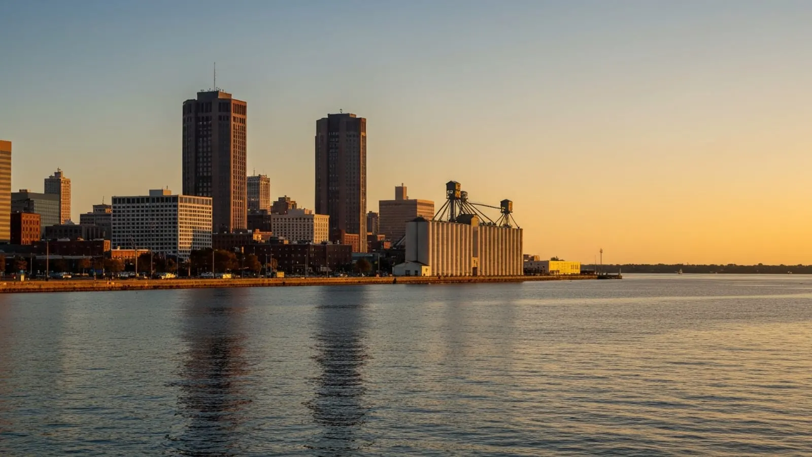 Editorial photograph of the downtown Buffalo skyline at golden hour viewed from the Lake Erie waterfront with historic grain elevators visible in the foreground.