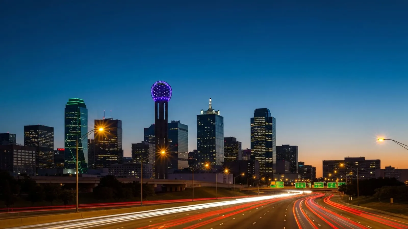 Dallas skyline at dusk featuring Reunion Tower and downtown high-rises with traffic light trails along Woodall Rodgers Freeway