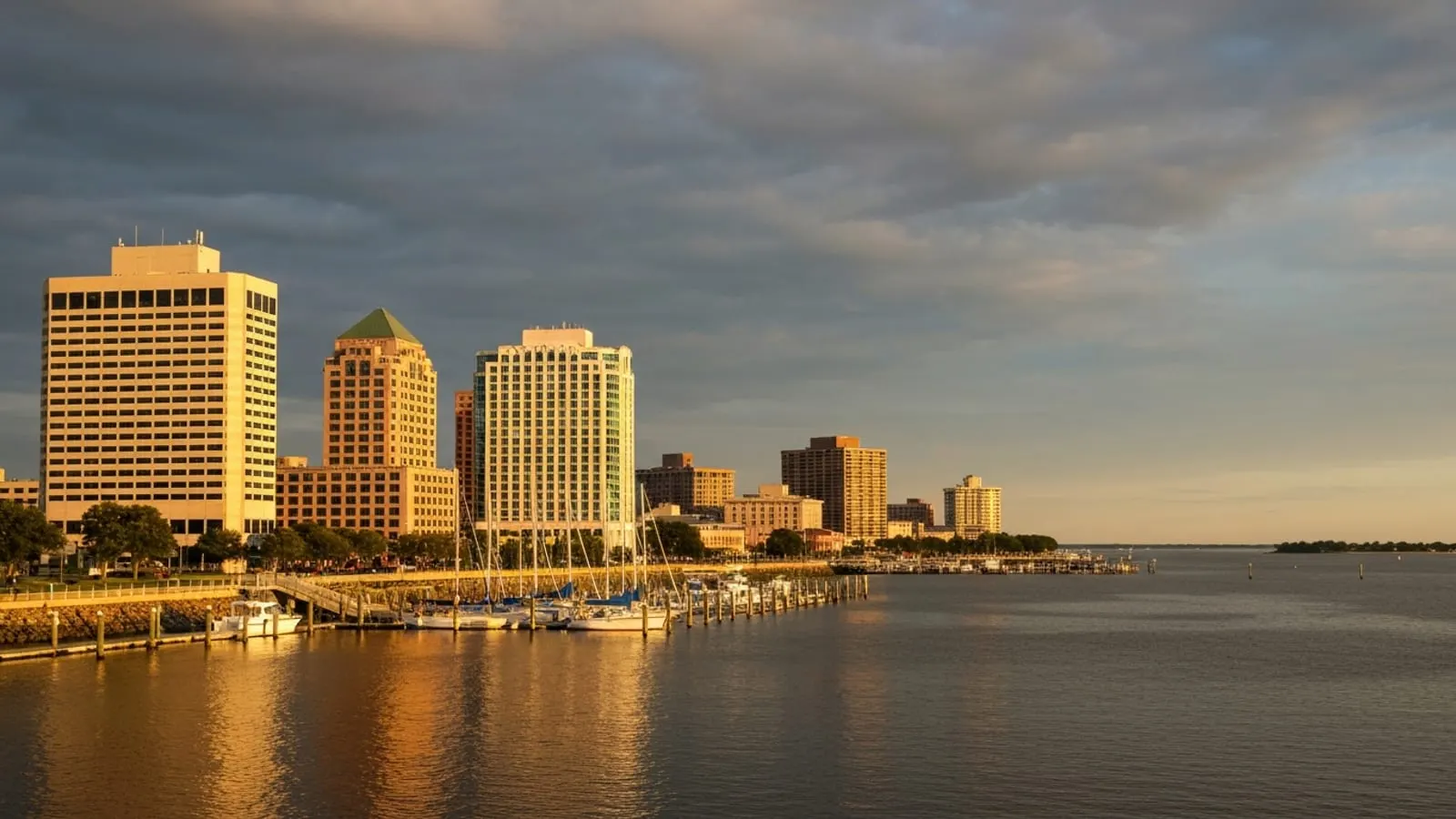 Editorial photograph of the Hampton Virginia waterfront and Hampton River at golden hour with mid-rise downtown buildings and a marina with sailboats.