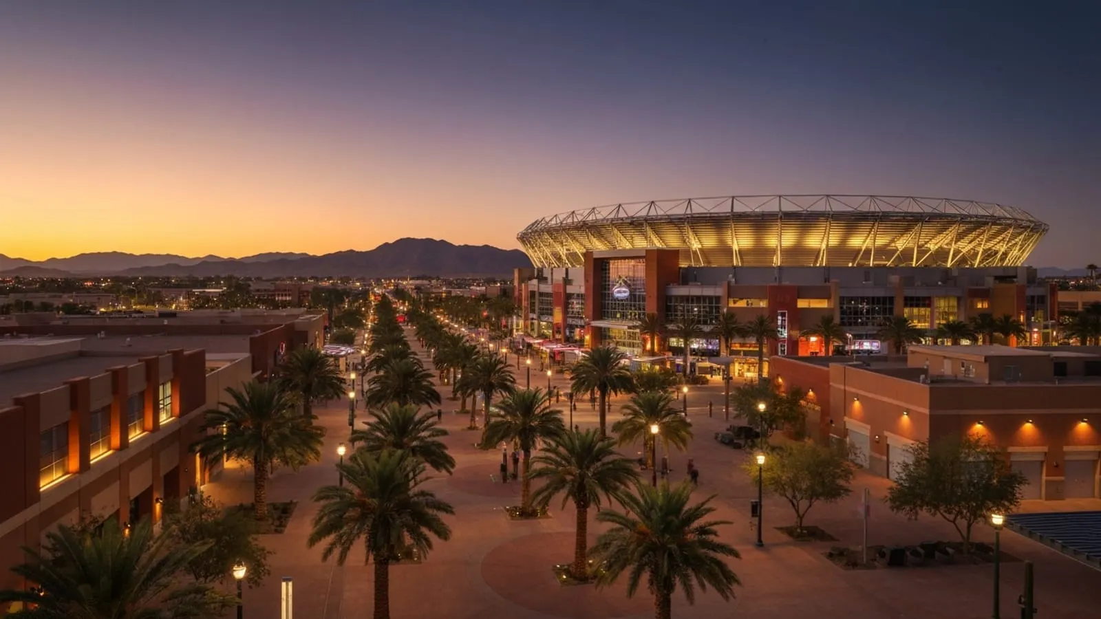 Editorial photograph of the Westgate entertainment district in Glendale Arizona at dusk with a large modern stadium structure illuminated and a wide pedestrian plaza with palm trees.