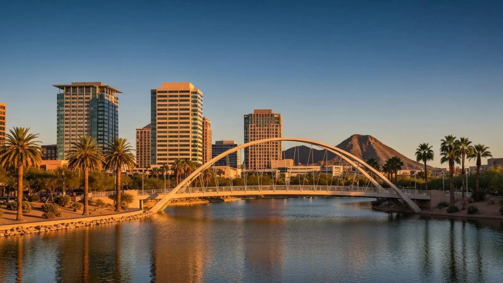 Editorial photograph of Tempe Town Lake at golden hour with the iconic pedestrian arch bridge, modern mid-rise downtown Tempe buildings in the background, and palm trees along the lakeshore.