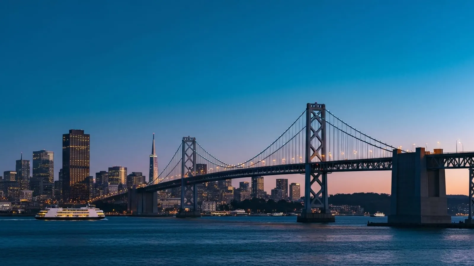 Editorial photograph of the San Francisco Bay Bridge at twilight with the downtown SF skyline including Salesforce Tower lit behind it.