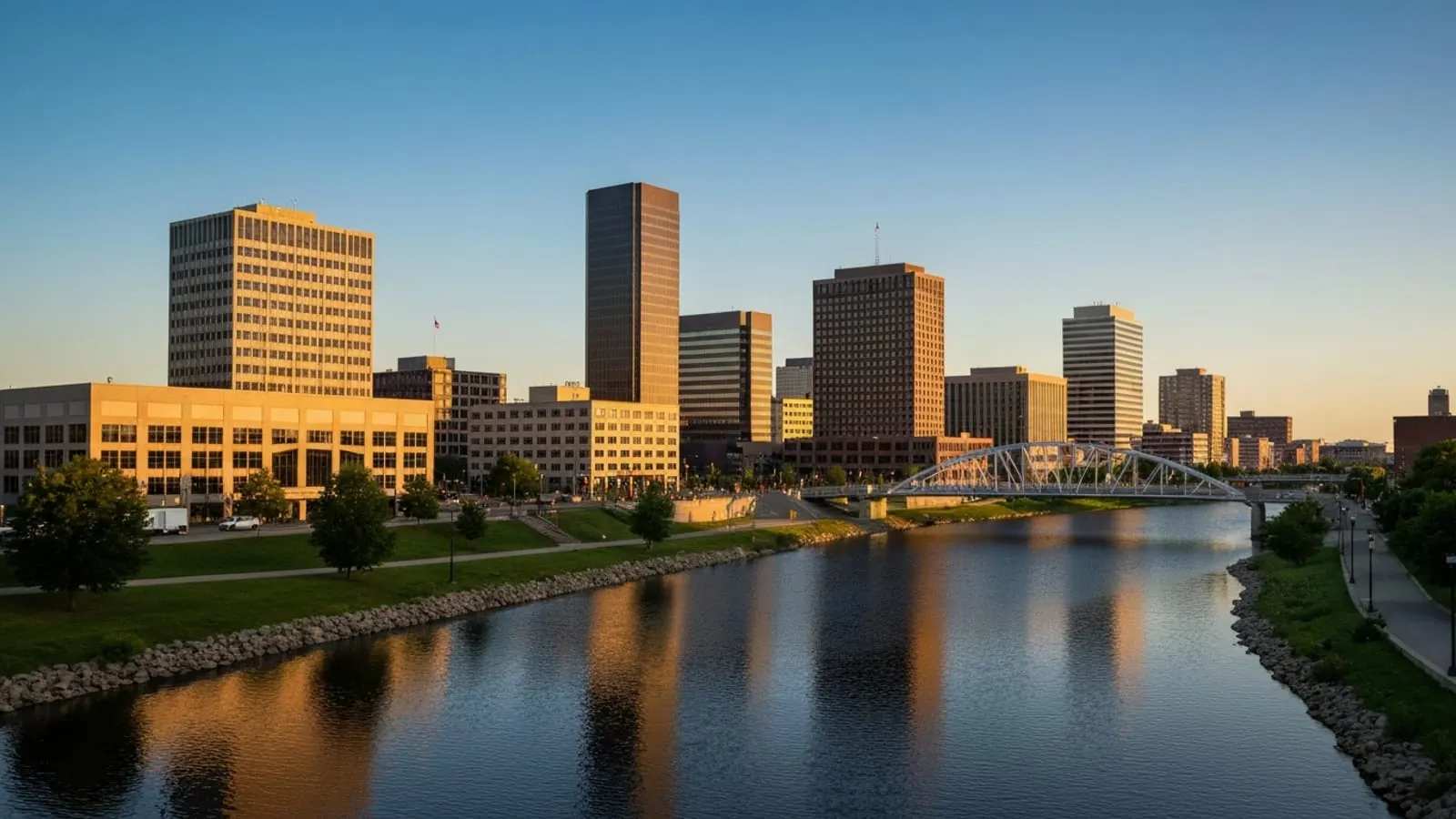 Editorial photograph of downtown Grand Rapids along the Grand River at golden hour with mid-rise downtown buildings reflected in the water.