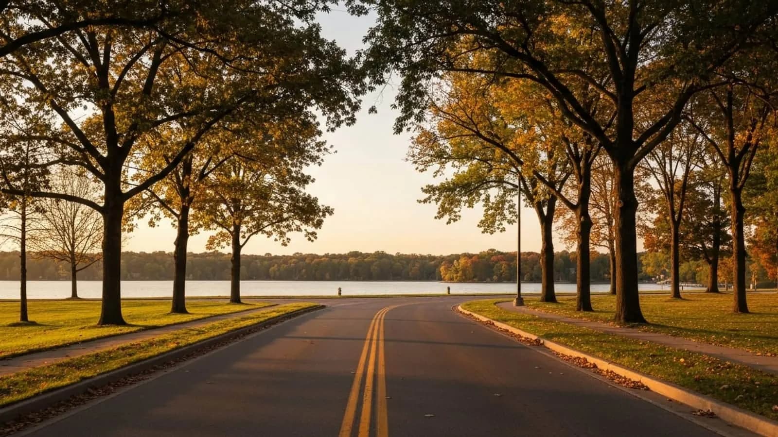 Editorial photograph of a tree-lined suburban Michigan parkway in Sterling Heights at golden hour with Stoney Creek Metropark forested shoreline in the background.
