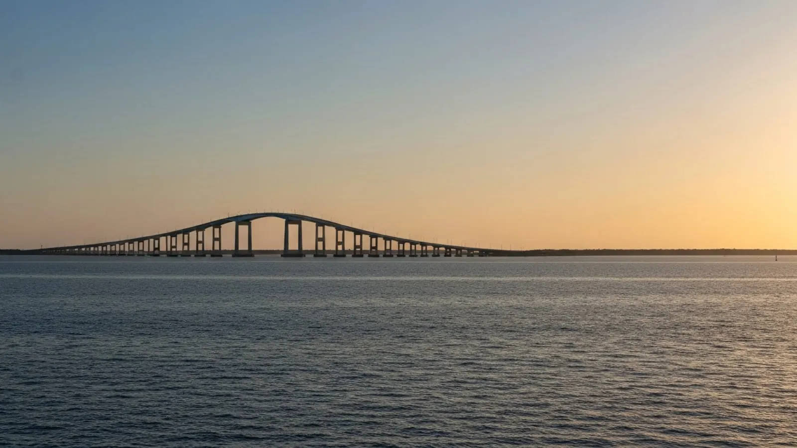 Editorial photograph of the Chesapeake Bay Bridge-Tunnel approach span stretching across broad bay waters at golden hour with dramatic Tidewater coastal light.