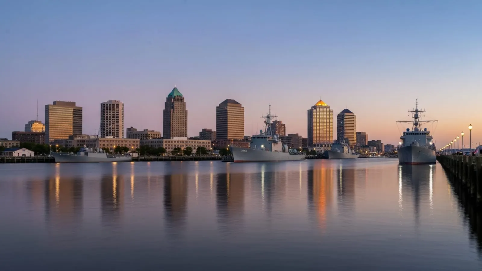 Editorial photograph of the Norfolk Virginia waterfront and Elizabeth River at golden hour with downtown skyline and naval ships docked in the distance.