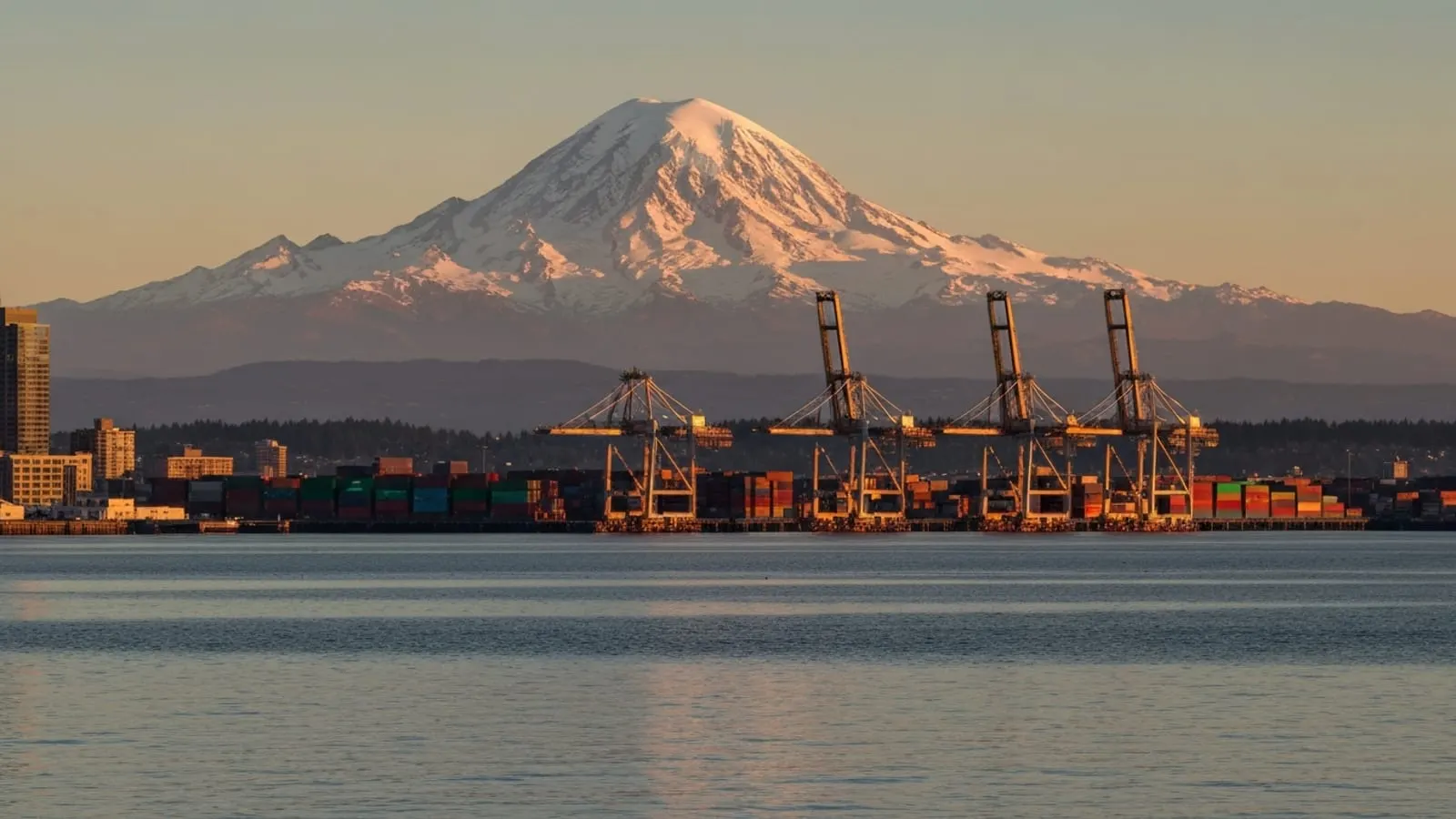 Editorial photograph of Mount Rainier rising massive and snow-capped above downtown Tacoma Washington at golden hour with the working Port of Tacoma container cranes silhouetted on Commencement Bay.